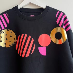 Può includere: Una felpa nera a girocollo con una stampa grafica di varie forme geometriche. Il design include cerchi con strisce rosa acceso, un cerchio dorato con una trama ondulata, un cerchio color corallo pieno, un semicerchio rosa acceso e un anello dorato.