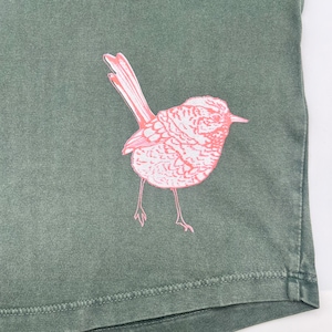 May include: A green t-shirt with a pink bird graphic printed on the front. The bird is facing to the right and has a long tail.
