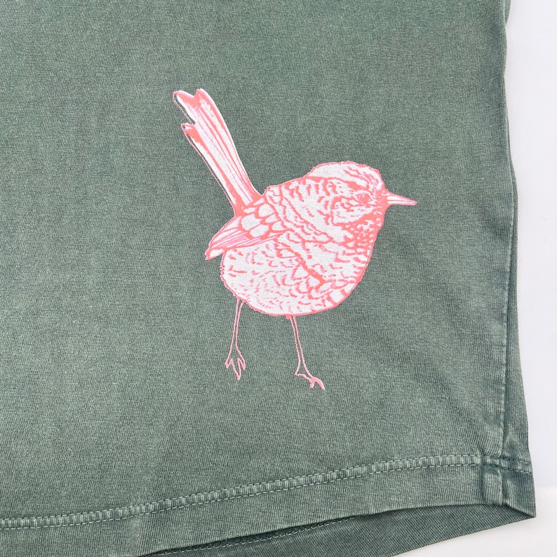 May include: A green t-shirt with a pink bird graphic printed on the front. The bird is facing to the right and has a long tail.