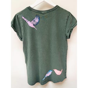 May include: A dark green t-shirt with a pink and blue bird design on the back. The birds are facing each other and are in a flying position.