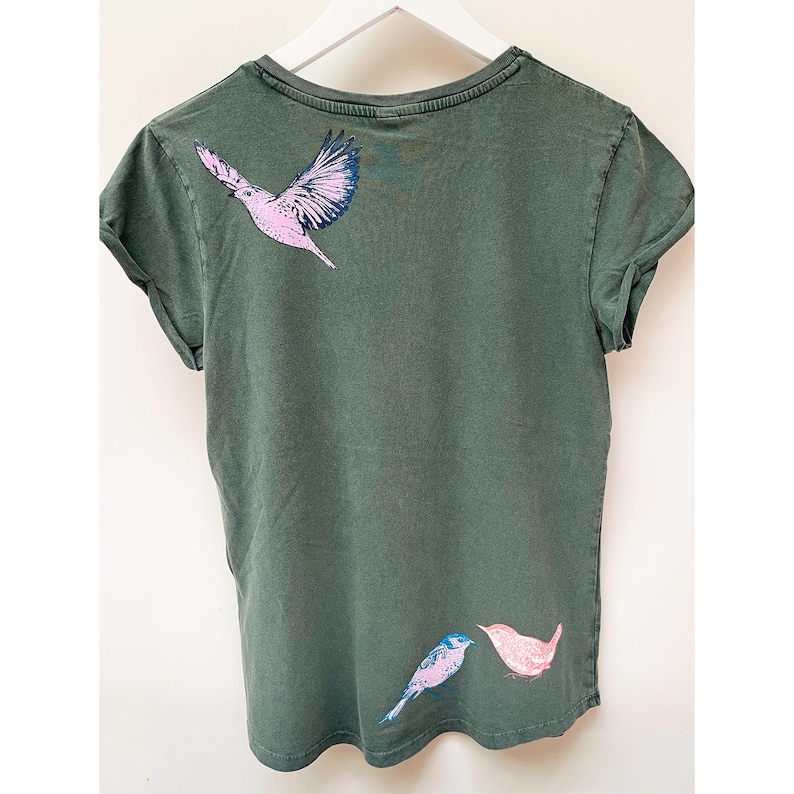 May include: A dark green t-shirt with a pink and blue bird design on the back. The birds are facing each other and are in a flying position.