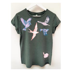 May include: A dark green t-shirt featuring a graphic print of five birds in flight. The birds are pink, blue and white.