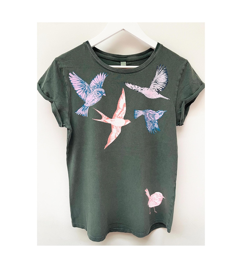 May include: A dark green t-shirt with a graphic print of five birds in flight. The birds are pink, blue, and white.