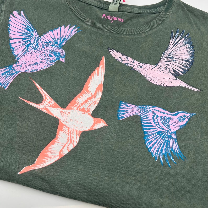 May include: A green t-shirt with a pink, blue, and orange bird print. The shirt has the text "Pinkijones" on the front.