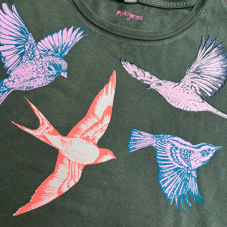 May include: A green t-shirt with a pink, blue, and red bird design. The text "Pinkijones" is printed on the shirt.