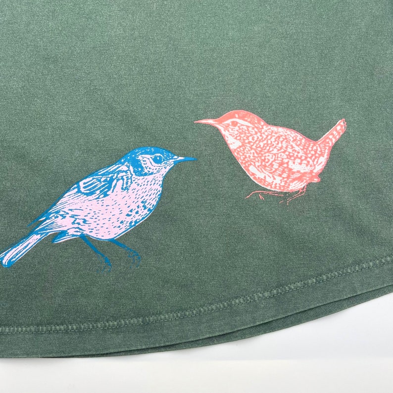 May include: A green t-shirt with a blue and pink bird design. The bird is facing left and has a long tail. The background is a dark green color.