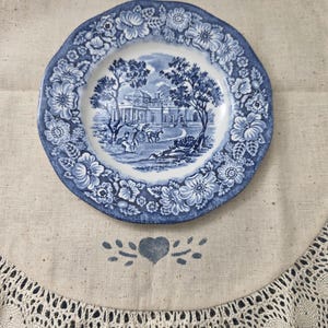 Può includere: Un piatto rotondo in ceramica con un motivo decorativo blu e bianco. Il centro raffigura un paesaggio con un grande edificio, alberi e una carrozza trainata da cavalli. Il bordo è decorato con una fitta cornice floreale di fiori.
