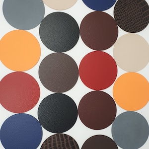 Könnte beinhalten: Sortiment von 16 runden Lederuntersetzern in verschiedenen Farben und Texturen, darunter Braun, Schwarz, Rot, Orange, Blau und Beige. Einige Untersetzer haben eine glatte Oberfläche, während andere eine strukturierte oder geprägte Oberfläche haben.