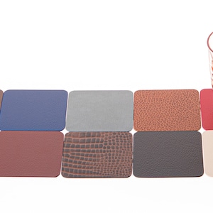 Może przedstawiać: Zestaw dziesięciu kwadratowych podkładek skórzanych w różnych kolorach i fakturach, w tym brązowy, niebieski, szary, pomarańczowy, zielony, czerwony i beżowy. Przezroczysty kieliszek z różowym odcieniem stoi na czerwonej podkładce.