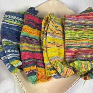 Könnte beinhalten: Vier Paar bunte Stricksocken werden auf einer cremefarbenen Oberfläche präsentiert. Die Socken weisen verschiedene Muster und Farbkombinationen auf, darunter Blau, Grün, Gelb, Orange und Rosa. Die Socken sind aus weichem, strukturiertem Garn gefertigt.