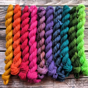 Könnte beinhalten: Eine Sammlung von zehn Garnknäuel in verschiedenen Farben, darunter Gelb, Orange, Rot, Rosa, Lila, Blau, Grün und Limettengrün. Das Garn ist in einer Reihe auf einer Holzoberfläche angeordnet.