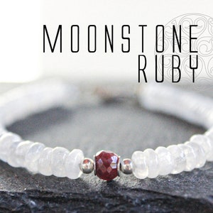 May include: A white moonstone bracelet with a faceted ruby gemstone and silver accents. The bracelet features the text "MOONSTONE RUBY". The bracelet is displayed on a dark grey surface.