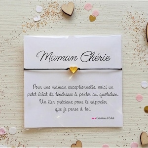 May include: A black string bracelet with a small gold heart charm is displayed on a white card. The card has the words "Maman Chérie" in script and a message in French. Wooden heart shapes and confetti surround the card.