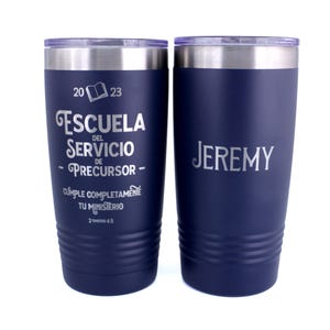 May include: Two dark blue stainless steel tumblers with clear lids. The left tumbler features the text 2023, an open book icon, ESCUELA DEL SERVICIO DE PRECURSOR, CUMPLE COMPLETAMENTE TU MINISTERIO, and 2 TIMOTEO 4:5. The right tumbler features the name JEREMY.