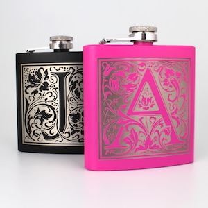 May include: Two hip flasks with engraved initials and floral designs. The flask on the left is black with a silver 'I' and the flask on the right is pink with a silver 'A'.