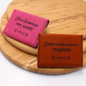May include: Two small, square, leather-like cases. One is hot pink with the text "FULLY ACCOMPLISH YOUR MINISTRY" and "2 TIM 4:5". The other is brown with the text "CUMPLE COMPLETAMENTE TU MINISTERIO" and "2 TIM 4:5".