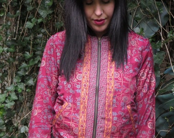 Könnte beinhalten: Burgunderfarbene Bomberjacke mit floralem Muster und einer zentralen vertikalen Bahn mit orangefarbenen und goldenen geometrischen Designs. Die Jacke hat einen schwarzen Reißverschluss, Bündchen und Saum. Lange Ärmel.