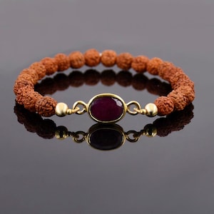 May include: A red gemstone bracelet with a gold setting. The bracelet is made of brown beads and has a gold clasp.