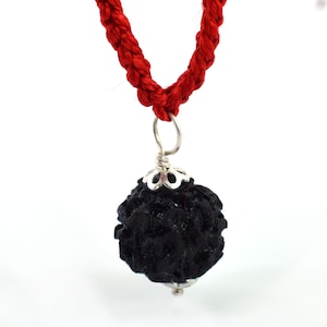 May include: A black rudraksha bead pendant on a red cord necklace. The bead is round and has a rough, textured surface. The pendant is attached to the cord with a silver bail.