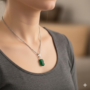 Peut inclure: Un collier en argent avec un petit pendentif en émeraude brute. L'émeraude est d'un vert vif et a une surface texturée. La chaîne est délicate et repose sur le cou d'une personne.
