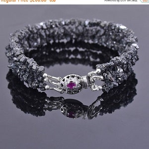 May include: A black diamond bracelet with a silver clasp featuring a small red gemstone. The bracelet is made of rough, uncut black diamonds.