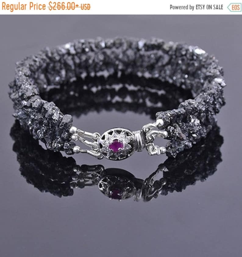 May include: A black diamond bracelet with a silver clasp featuring a small red gemstone. The bracelet is made of rough, uncut black diamonds.