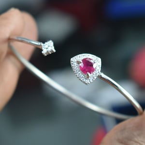 May include: A silver bracelet featuring a triangular, ruby-red gemstone encircled by small, clear stones. The open-ended design includes a cluster of clear stones on the opposing side. The bracelet is a piece of jewellery.