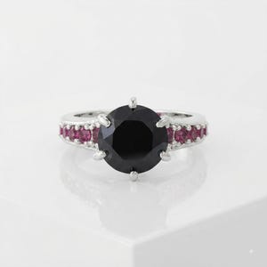 May include: A silver ring featuring a large, round, black center stone held by six prongs. The band is accented with a row of small, round, vibrant pink stones set along each side, creating a contrast against the polished silver metal finish.