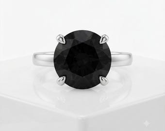 May include: A ring featuring a large, round-cut black gemstone set in a polished silver-toned metal band. The stone is secured by four prominent prongs, creating a clean and symmetrical design that highlights the contrast between the dark center.