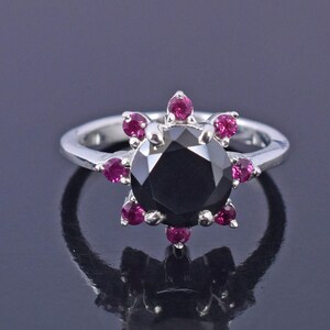 May include: A silver ring featuring a large, round black gemstone at the center, surrounded by smaller, vibrant pink gemstones. The ring's design is reminiscent of a flower, with the pink stones forming the petals. The band is a polished silver.