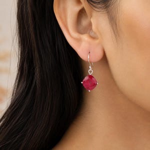 Peut inclure: Une boucle d'oreille pendante en argent avec une pierre précieuse rouge foncé taillée en coussin à facettes. La pierre est fixée dans un sertissage en argent à quatre griffes, suspendue à un crochet simple, formant un bijou classique.