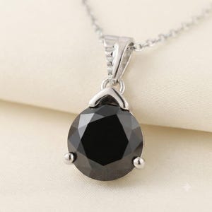 Peut inclure: Un collier pendentif en argent avec une grande pierre noire ronde à facettes, maintenue par un sertissage à trois griffes. Le pendentif est fixé à une fine chaîne en argent, présentant une finition métallique polie et un design symétrique.