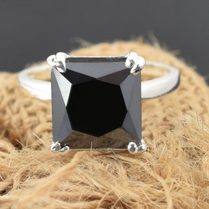Peut inclure: Une bague en argent avec une grande pierre noire de forme carrée. La bague est posée sur une surface brune texturée.