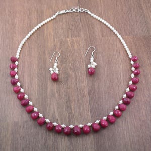May include: A necklace and earring set featuring a silver chain, small white pearls, and vibrant red teardrop-shaped beads. The earrings mirror the necklace's design, creating a coordinated jewelry set. The set is displayed on a wooden surface.