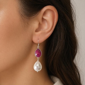 Può includere: Orecchini pendenti con una gemma rosa scuro sfaccettata a forma di goccia sopra una grande perla bianca a forma di pera. Gli orecchini sono appesi a un gancio d'argento. I gioielli sono esposti vicino all'orecchio di una persona.