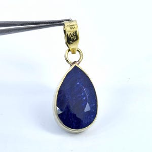 May include: A teardrop-shaped pendant with a faceted, deep blue gemstone. The gem is set in a gold-colored metal frame. The pendant is held by tweezers against a white background. The top of the pendant has a small loop for a chain.