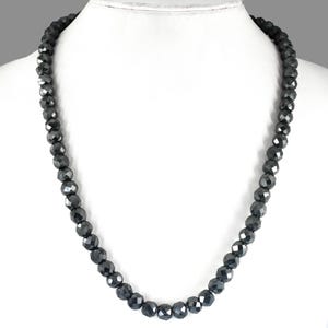 Può includere: Una collana di perline nere sfaccettate, esposta su un manichino bianco. Le perline sono di dimensioni uniformi e infilate insieme, creando un design simmetrico. La collana è di colore antracite scuro e le perline hanno una qualità riflettente.