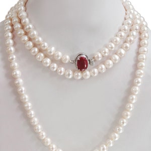 Pode incluir: Um colar de pérolas brancas com um fecho de pedra vermelha. O colar é de duas voltas e tem um design clássico.