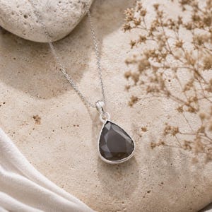 Peut inclure: Un collier en argent avec un pendentif en forme de goutte d'eau orné d'une pierre gris foncé à facettes. Le pendentif est suspendu à une fine chaîne en argent, présentant une monture métallique polie qui encadre la pierre précieuse sombre.