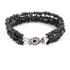 May include: A black diamond bracelet with a silver clasp featuring a small red gemstone. The bracelet is made of multiple strands of black diamond beads.