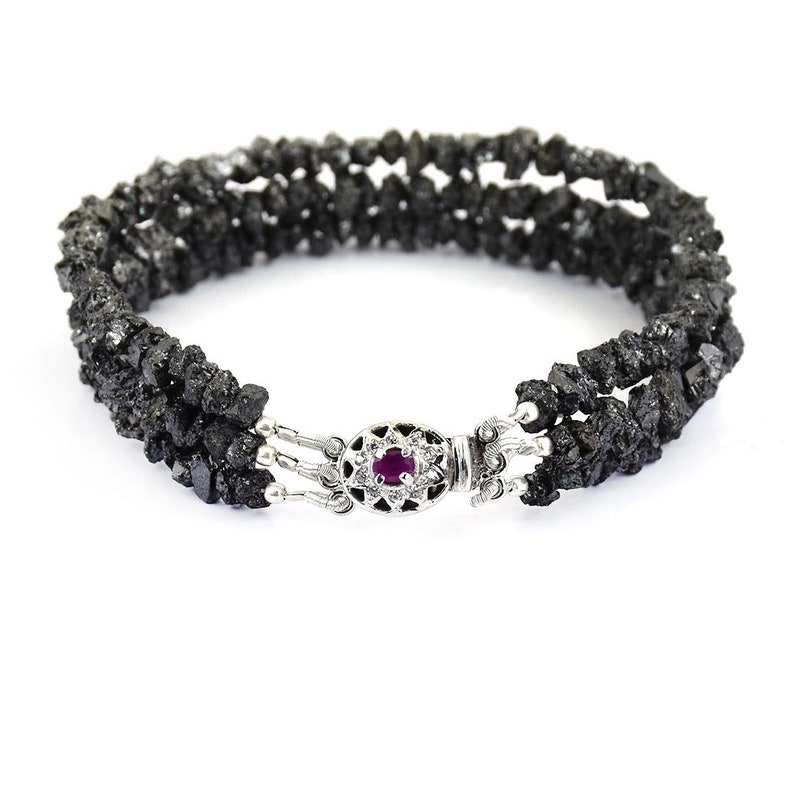 May include: A black diamond bracelet with a silver clasp featuring a small red gemstone. The bracelet is made of multiple strands of black diamond beads.