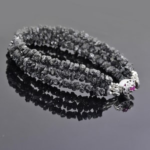 May include: A black diamond bracelet with a silver clasp and a small red gemstone. The bracelet is made of multiple strands of rough black diamonds.
