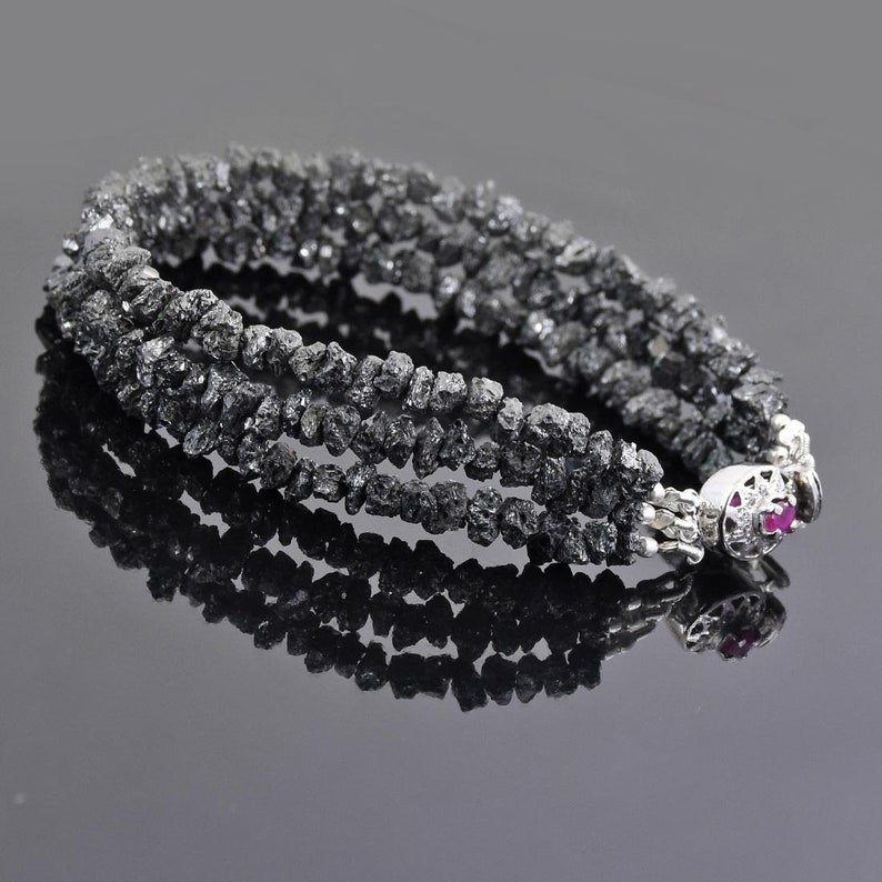 May include: A black diamond bracelet with a silver clasp and a small red gemstone. The bracelet is made of multiple strands of rough black diamonds.