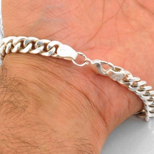 May include: A silver curb chain bracelet with a lobster clasp closure. The bracelet is shown on a person's wrist.