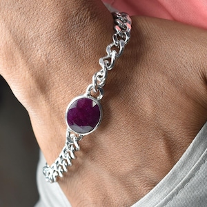 May include: A silver bracelet with a large, round, deep red gemstone at the center. The bracelet has a chain-link design. The gemstone is set in a silver bezel. The bracelet is on a person's wrist.