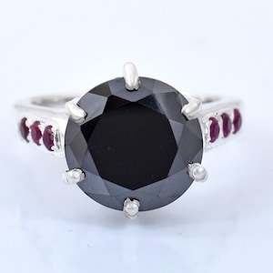 Peut inclure: Bague en argent ornée d'une grosse pierre noire facettée en son centre. La bague est ornée de petites pierres rouges rondes sur le pourtour. L'ensemble est présenté sur fond blanc.