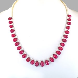 Op de afbeelding: Een ketting met een streng kleine, witte parels afgewisseld met levendige, druppelvormige, gefacetteerde rode edelstenen. De ketting wordt gepresenteerd tegen een witte achtergrond, die het contrast tussen de kleuren en texturen benadrukt.