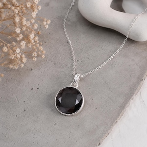 Peut inclure: Un collier en argent avec un pendentif rond en pierre noire à facettes, serti dans une monture en argent simple. Le pendentif est suspendu à une fine chaîne en argent, mettant en valeur une finition polie et une surface sombre et réfléchissante.