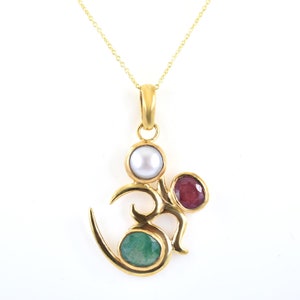May include: Gold pendant necklace featuring an Om symbol with three gemstones: a white pearl, a red ruby, and a green emerald.