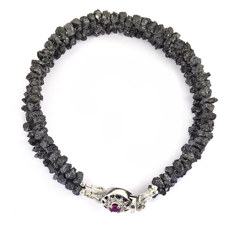 May include: A black diamond bracelet with a silver clasp featuring a small red gemstone. The bracelet is made of rough, uncut black diamonds.
